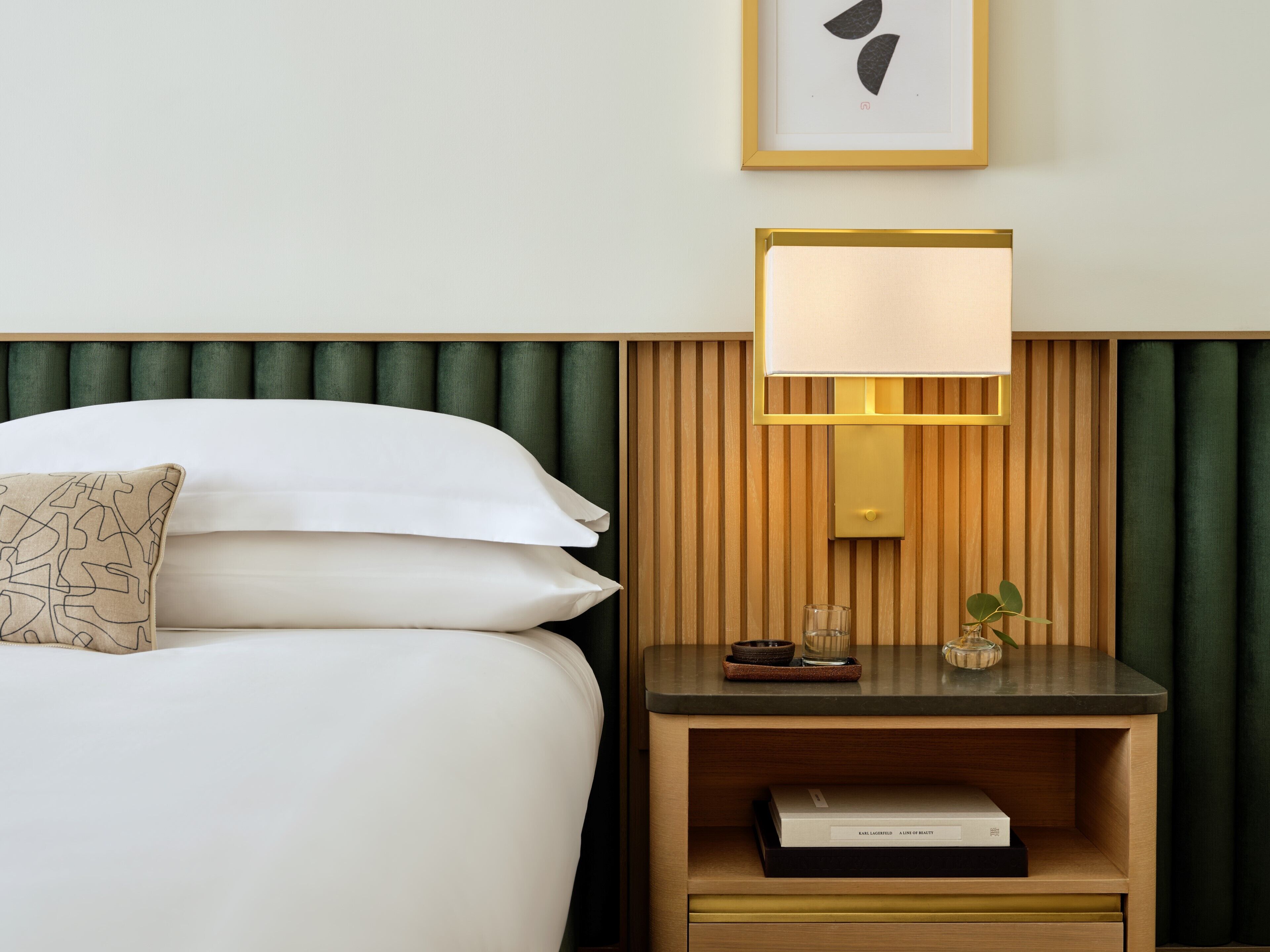 A neatly made bed with pillows, a bedside lamp, a small nightstand with a book, and a framed picture on the wall.