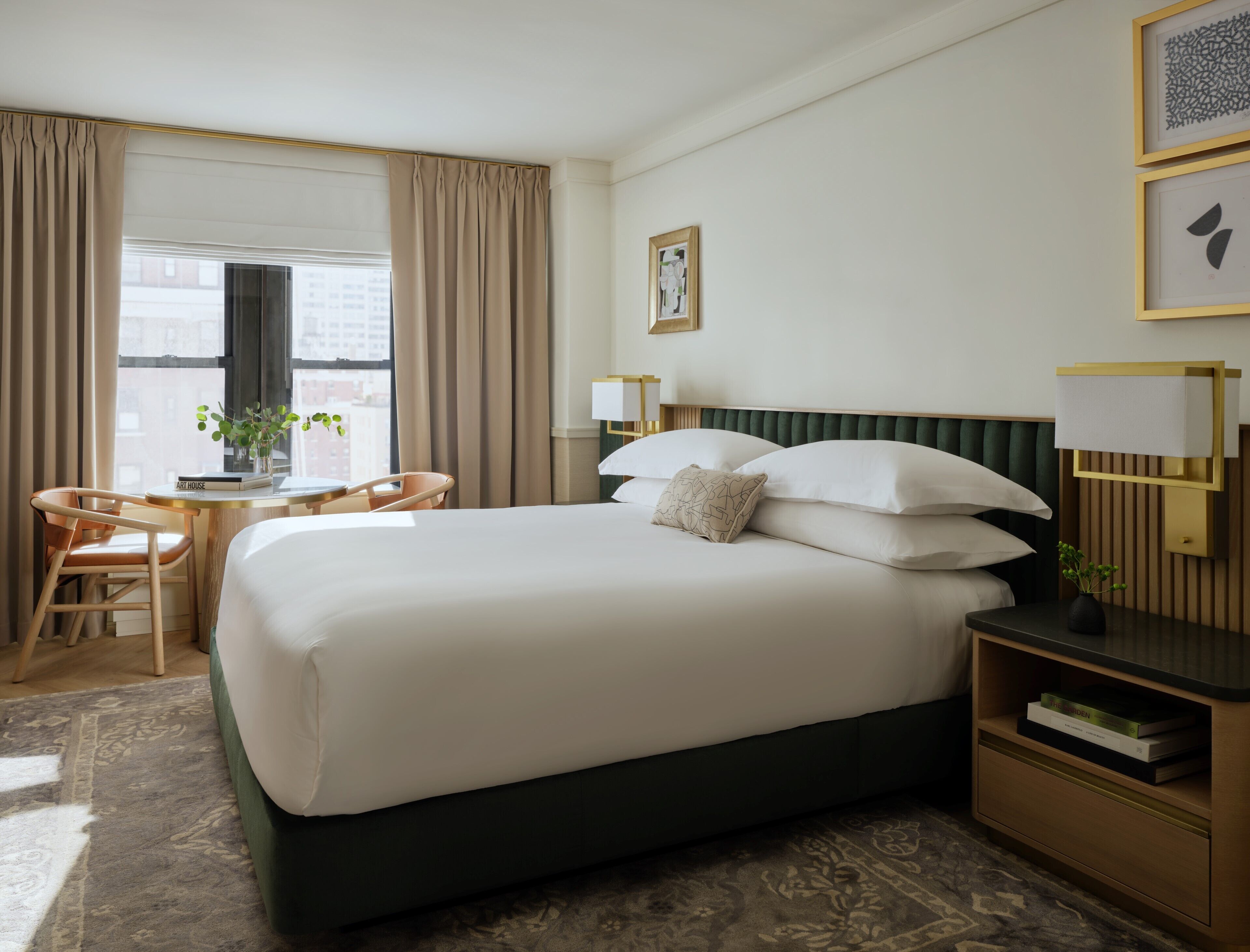 A hotel room with a large bed, a desk with a chair, a window with curtains, and a bedside table with a lamp.