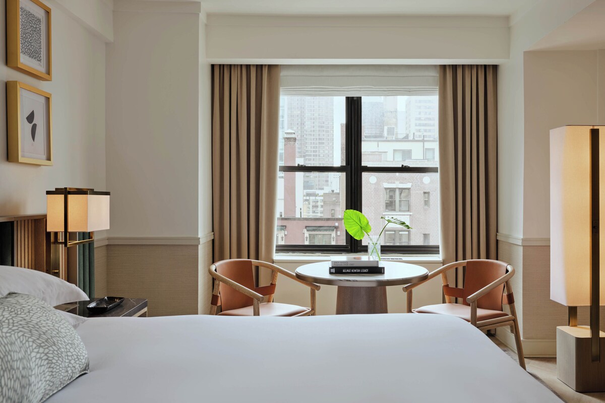A hotel room with a bed, two chairs, a small table with books, a window with curtains, and a lamp.
