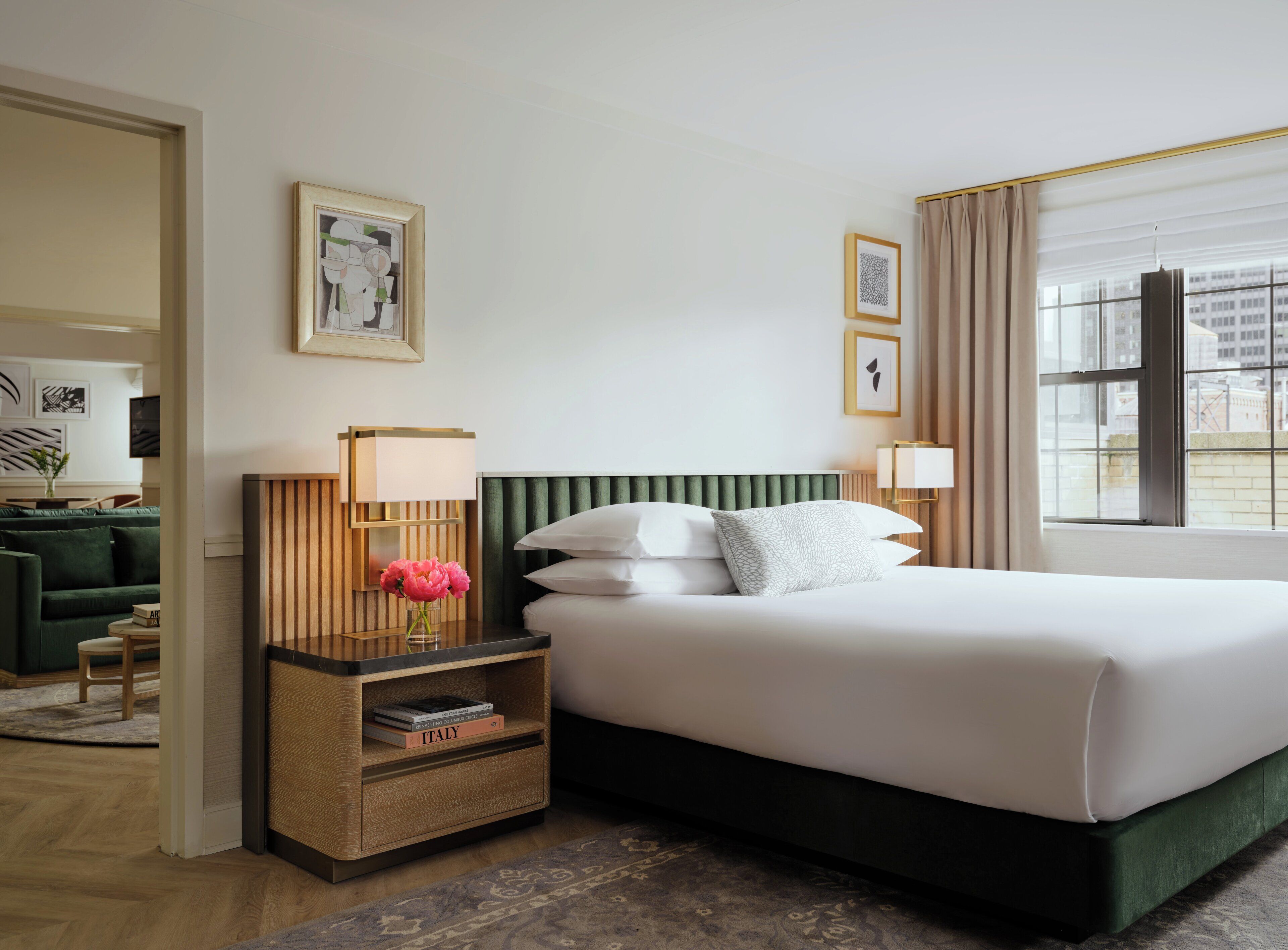 A hotel room with a bed, bedside tables, a green headboard, and a view of the city through large windows.