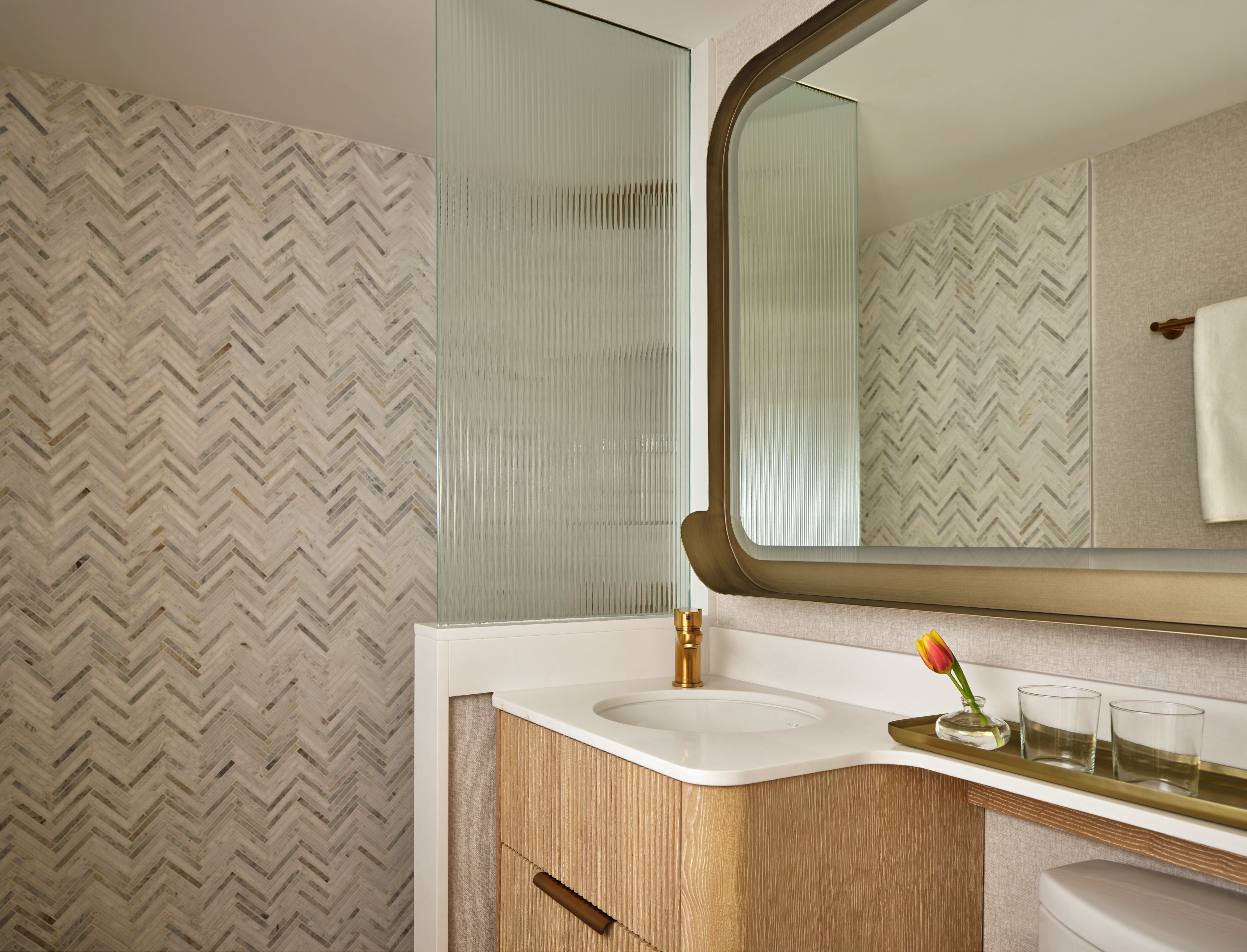 A modern bathroom with a herringbone patterned wall, a white sink, and a mirror with a gold frame.