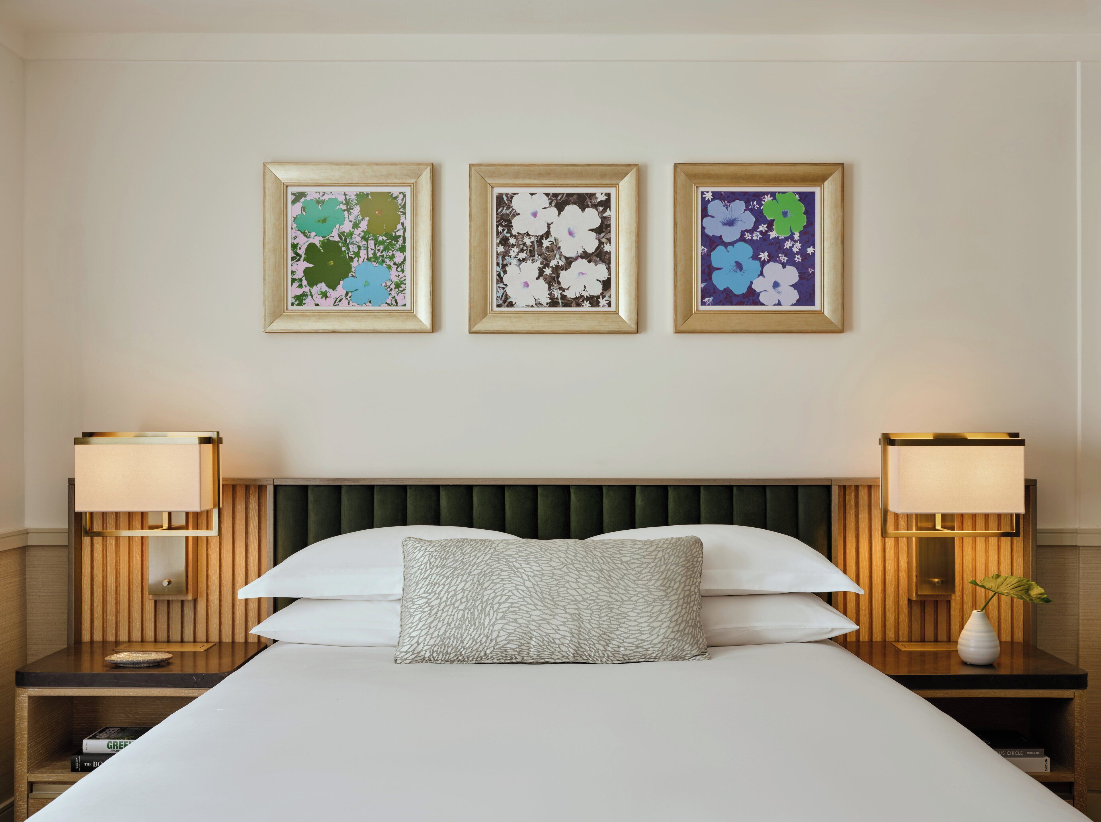 A hotel room with a bed, two bedside tables, and three framed floral artworks on the wall.