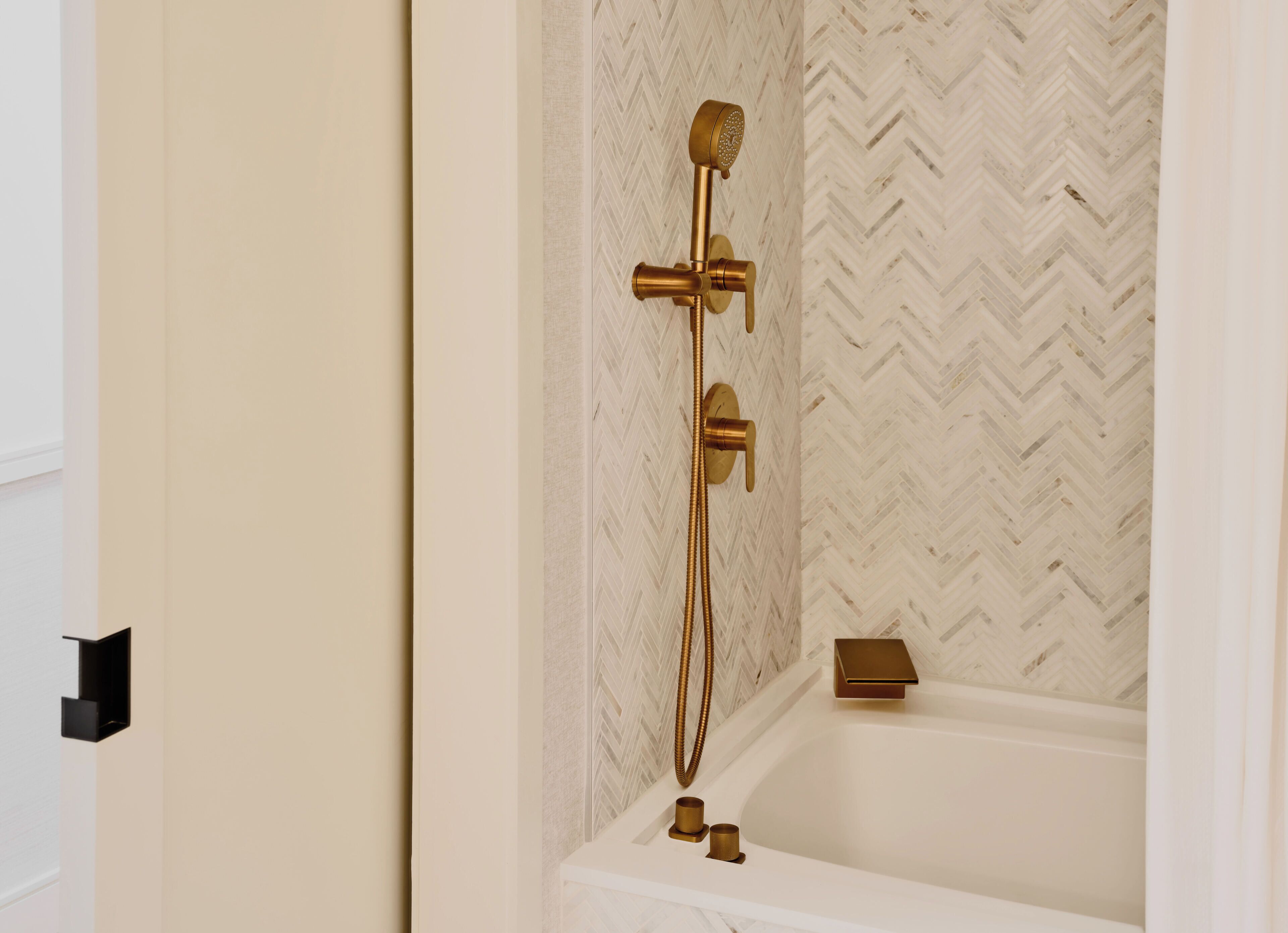 A bathroom with a herringbone patterned wall, a gold showerhead, and a white bathtub.