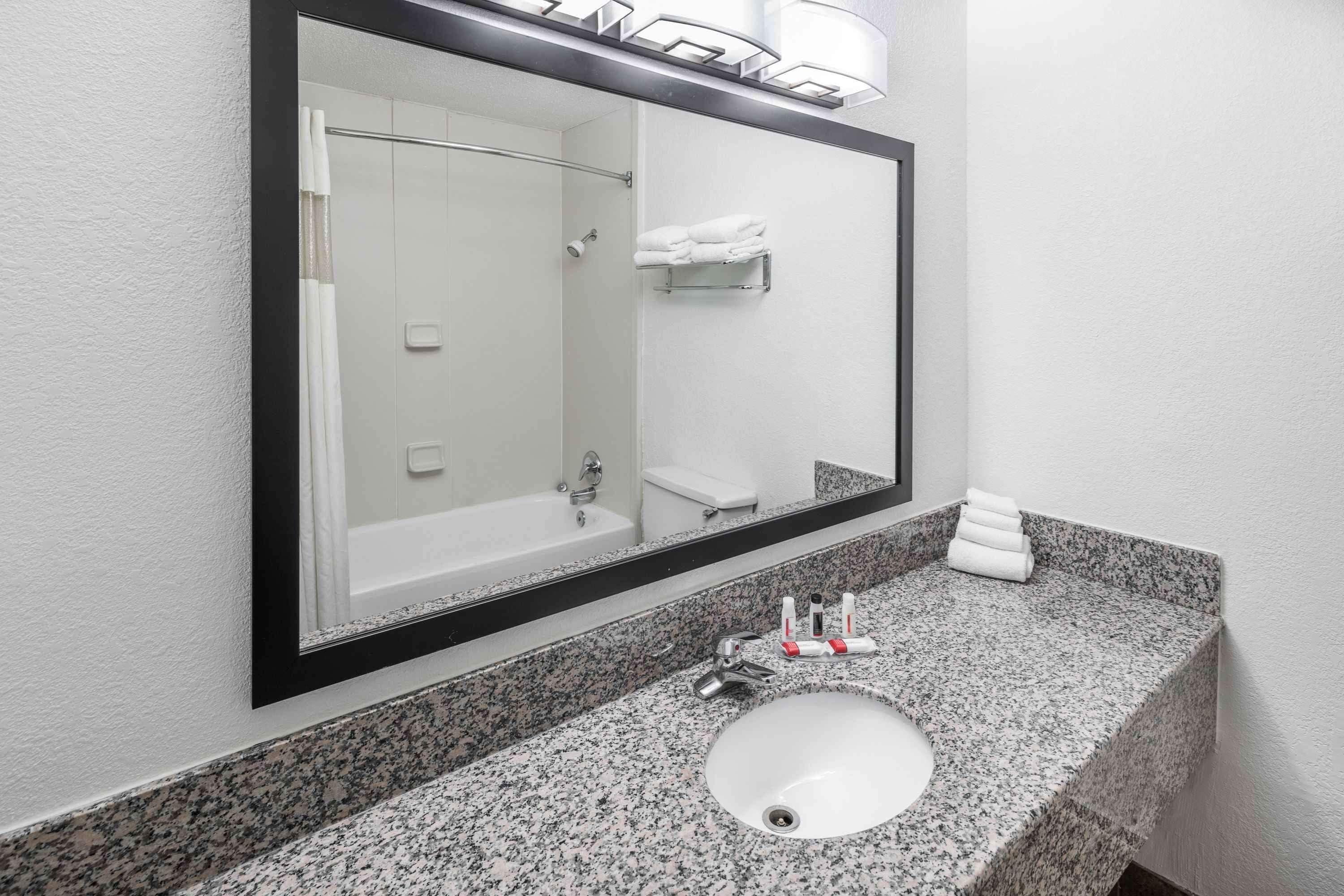A bathroom with a granite countertop, a white sink, a mirror, a towel rack, and a shower visible in the reflection.