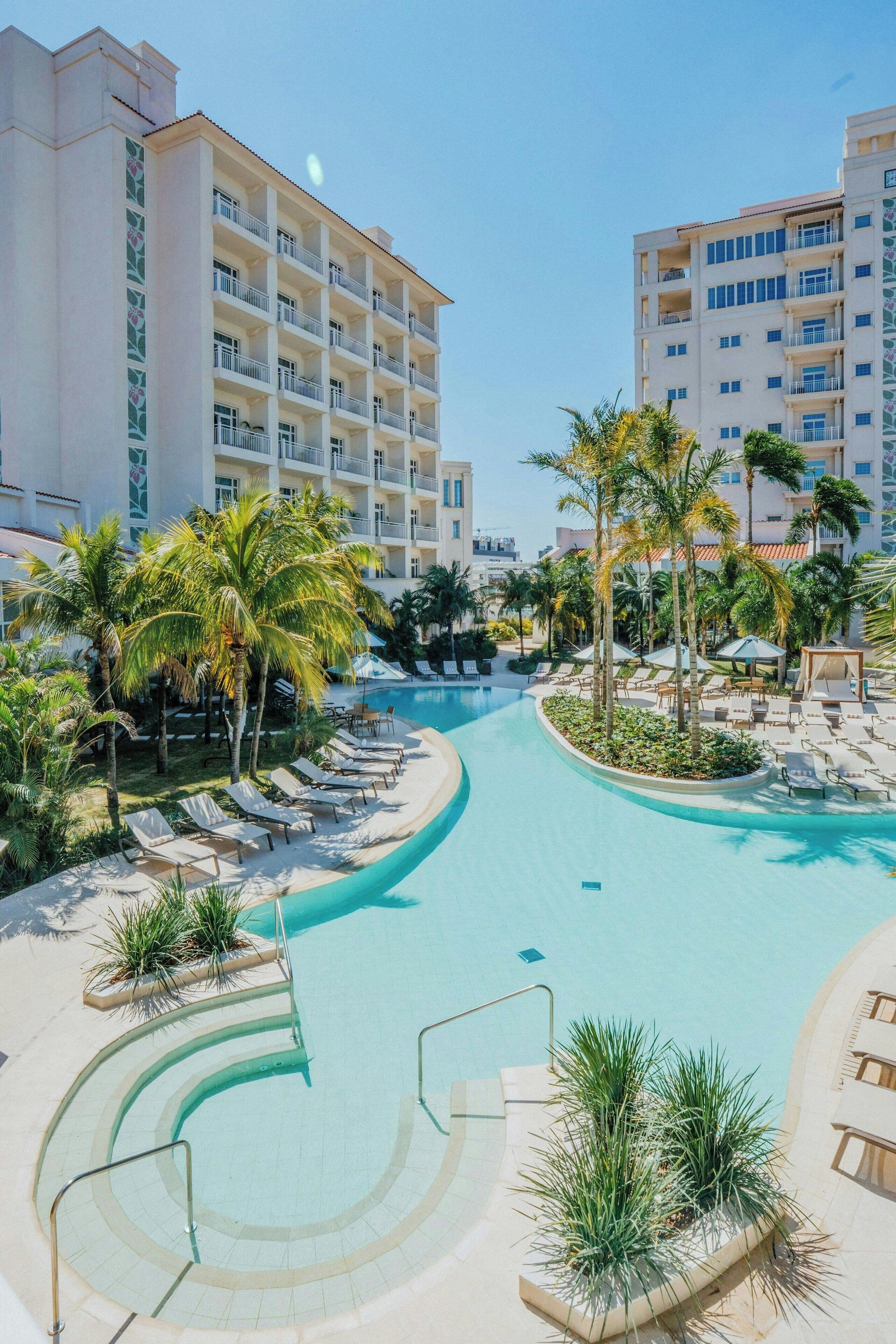 Zona de piscina en un resort con sillones de descanso y palmeras, rodeada de edificios de varios pisos.