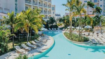 Zona de piscina en un resort con sillones de descanso y palmeras, rodeada de edificios de varios pisos.