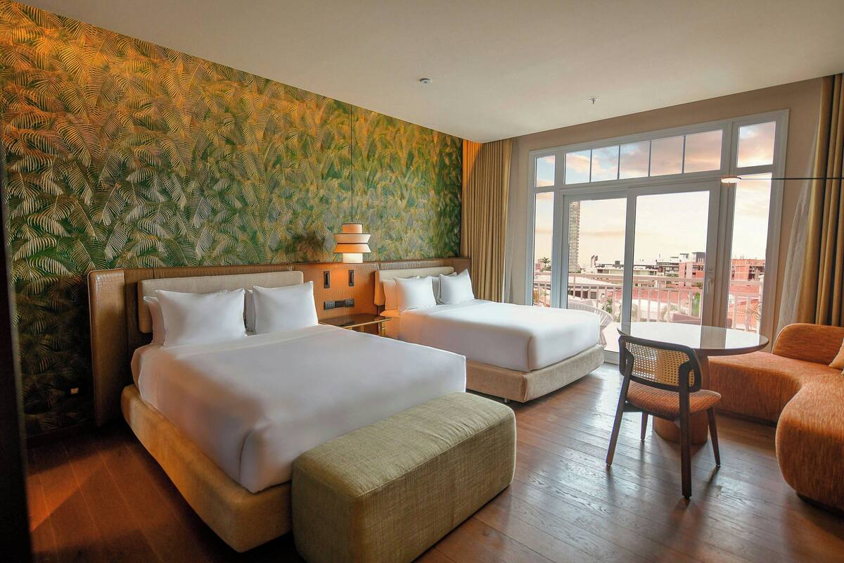 A hotel room with two beds, a sitting area with a chair, a small table, and a large window offering a city view.