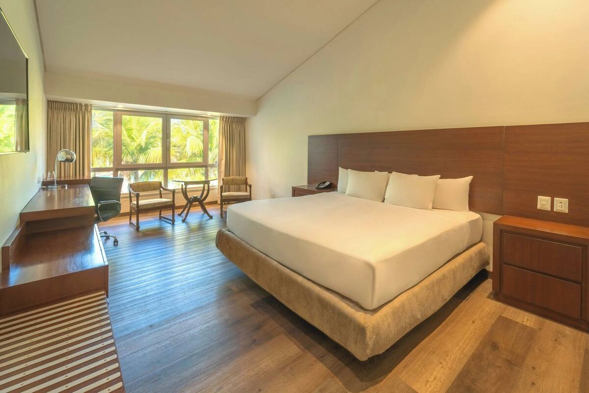 A hotel room with a large bed, wooden flooring, a desk with a chair, a small table, and a view of palm trees through the windows.