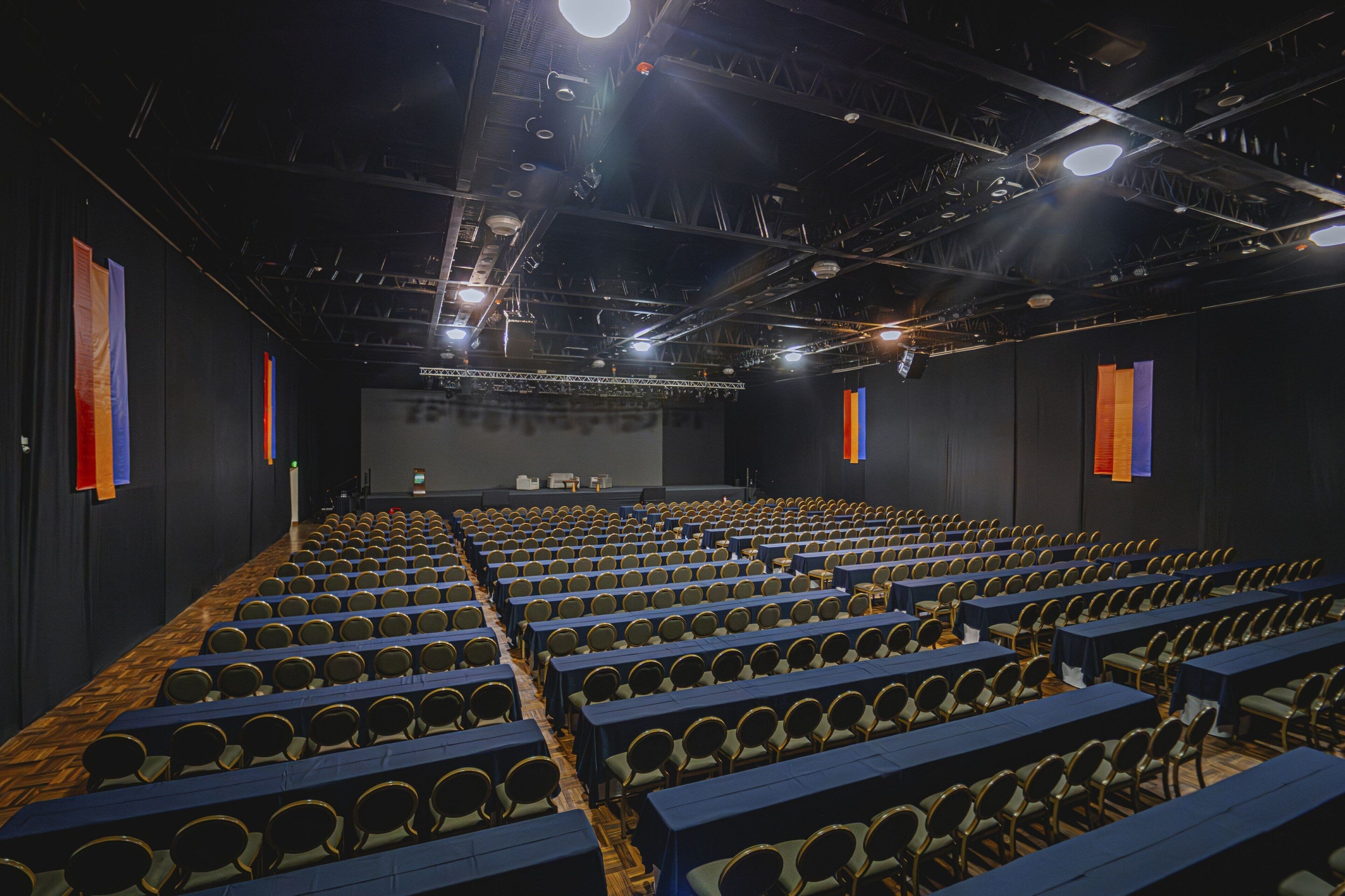 Uma ampla sala de conferências com fileiras de cadeiras e mesas, um palco na frente e um teto escuro com luminárias.
