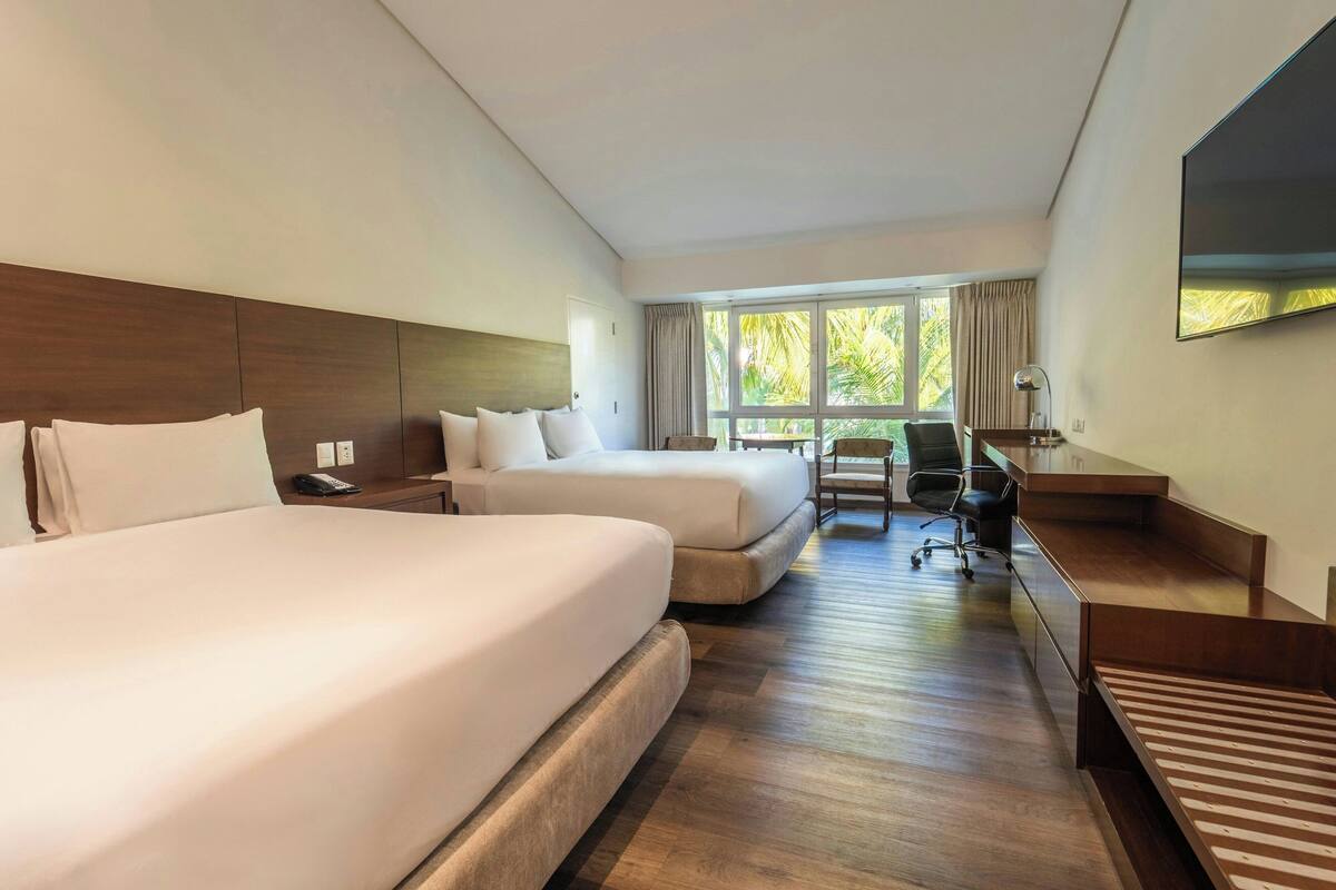A hotel room with two beds, a desk, a chair, a TV, and a large window with curtains.