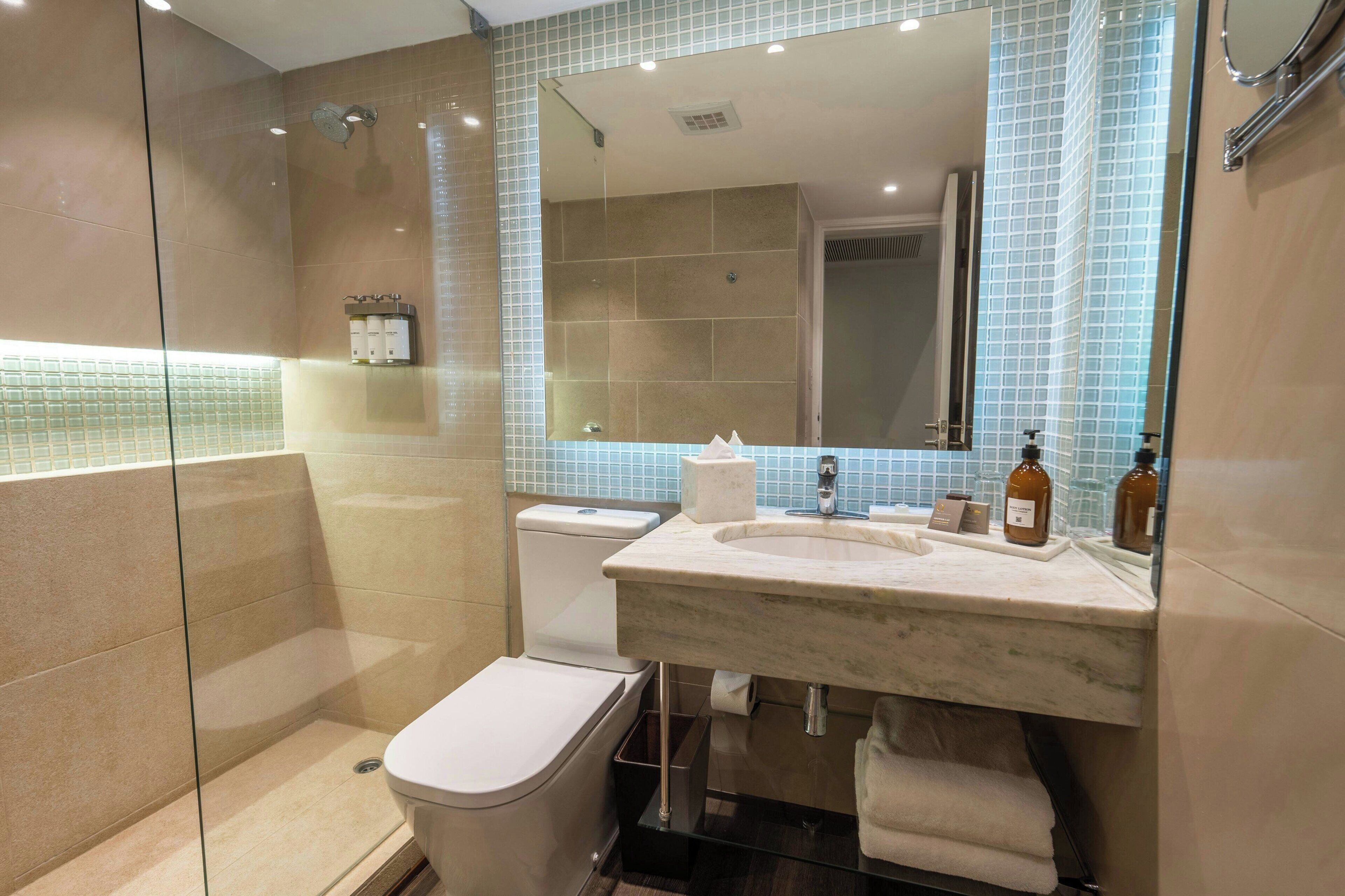 Un baño moderno con ducha de vidrio, inodoro blanco y lavamanos de mármol con artículos de aseo.