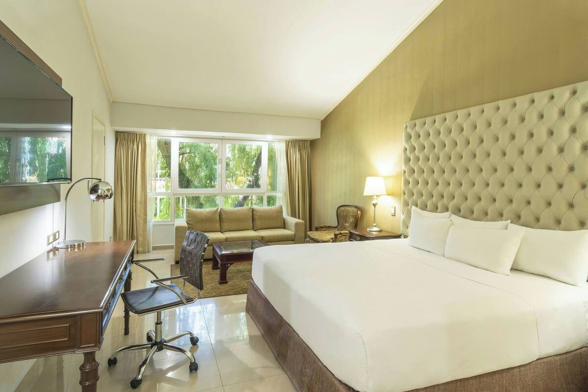 A hotel room with a large bed, a desk, a chair, a sofa, and a television.