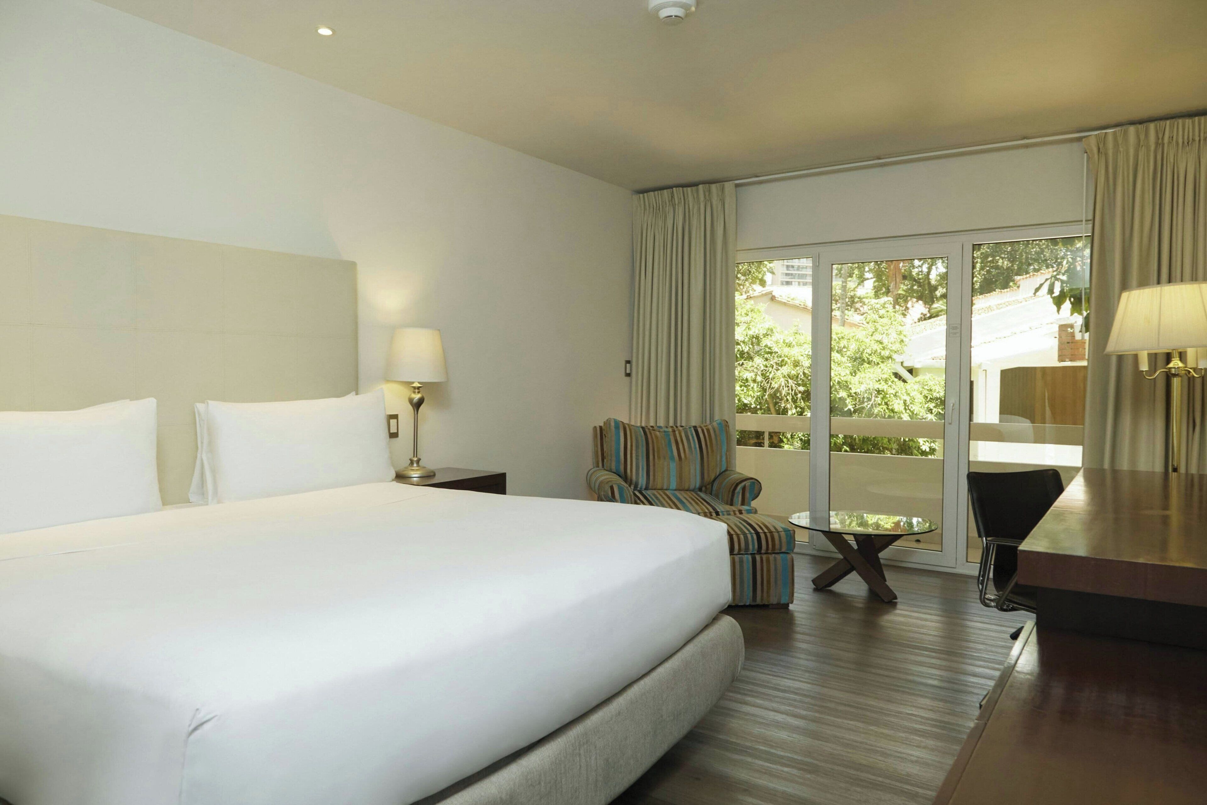 Habitación de hotel con una cama grande, un sillón pequeño, una mesita de vidrio y vistas a zonas verdes a través de un ventanal corredero.