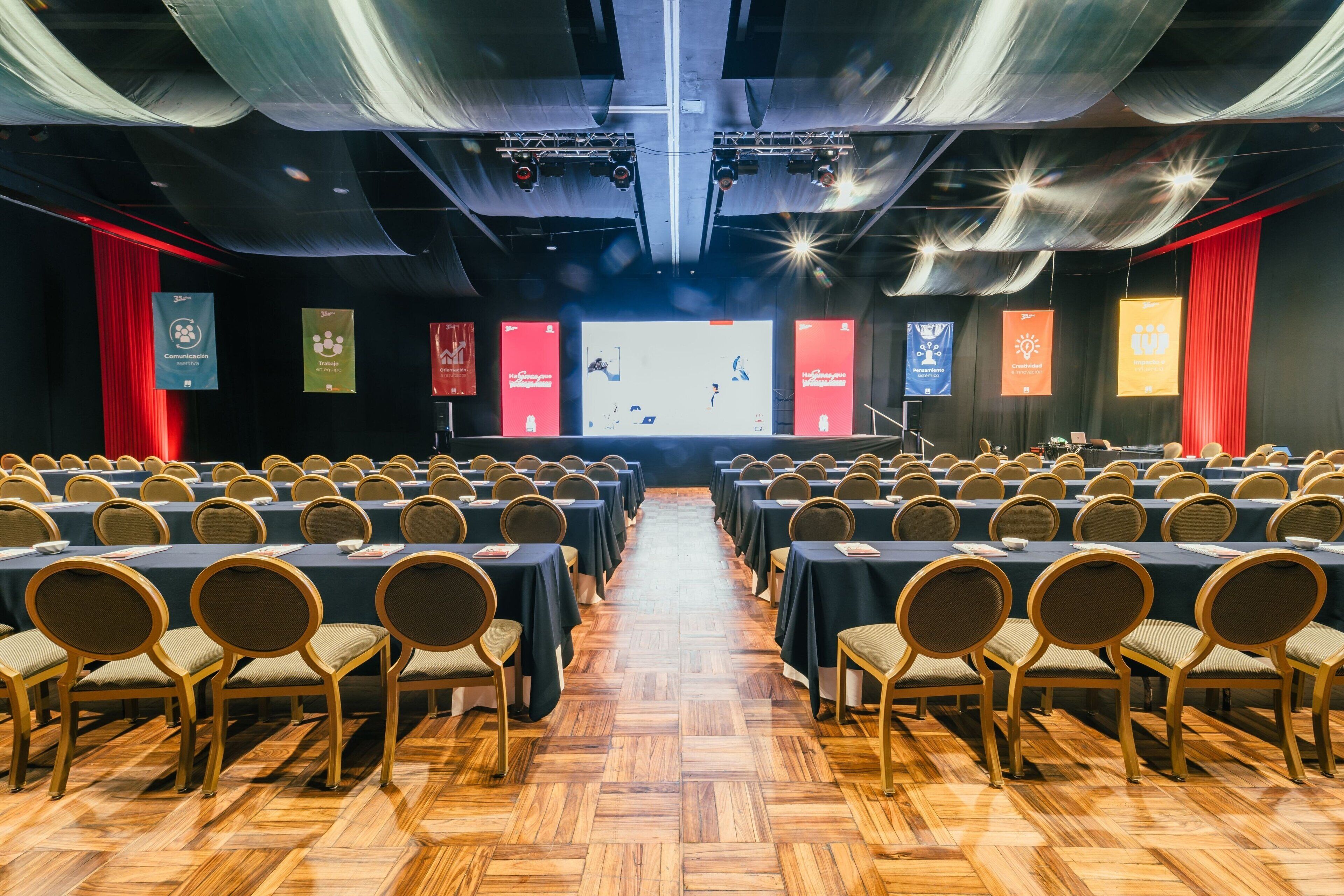 A conference room with rows of tables and chairs, a stage with banners, and a wooden floor.