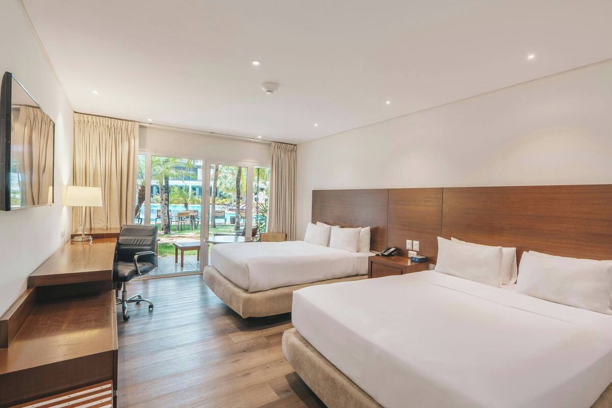 A hotel room with two beds, a desk, a chair, a lamp, and a view of the outdoors.