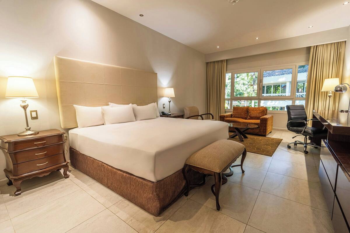 A hotel room with a large bed, a desk, a chair, a lamp, and a view of the outdoors.