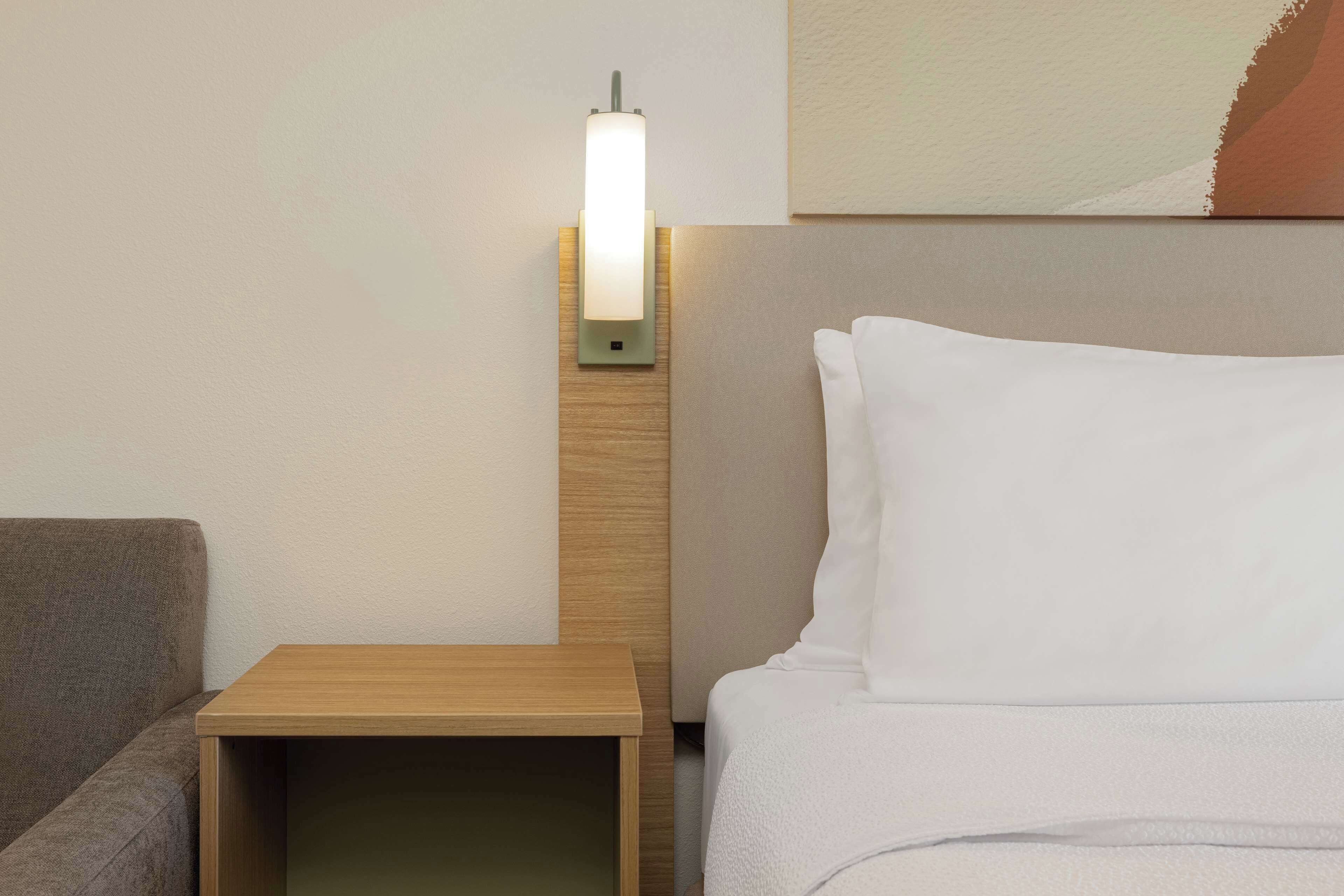 Habitación de hotel impecable con mesita de noche de madera, cama con ropa blanca y lámpara de pared.