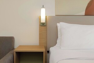 Habitación de hotel impecable con mesita de noche de madera, cama con ropa blanca y lámpara de pared.