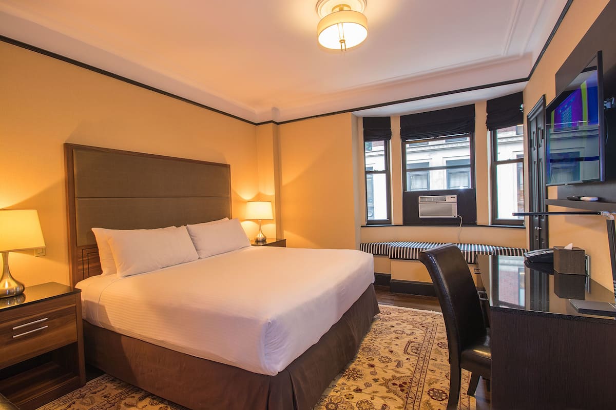 Hotellihuone, jossa on suuri sänky, yöpöydällä lamput, työpöytä tuolilla, televisio ja ikkuna, jossa on sälekaihtimet.