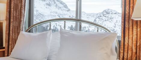 A hotel room with a bed, a window with curtains, and a view of snow-covered mountains.