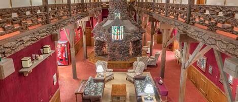 A high-ceilinged hall with a central stone fireplace, wooden railings, and a red carpeted floor.