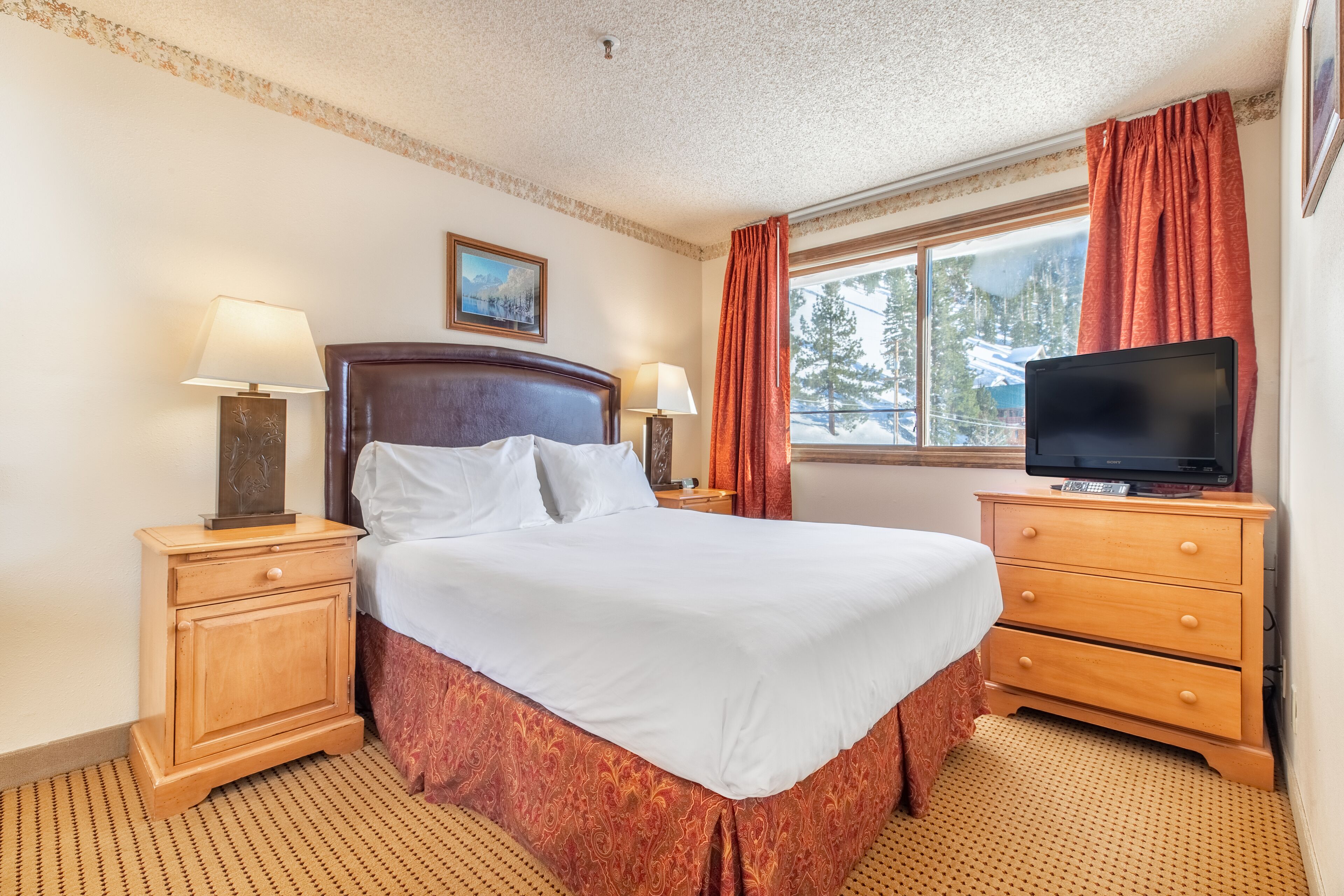 A neatly made bed with white linens, a wooden nightstand with a lamp, a wooden dresser with a television, and a window with curtains.