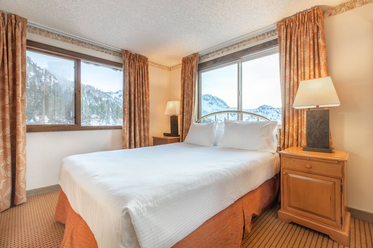 Ein Hotelzimmer mit einem großen Bett, zwei Nachttischen, einer Lampe und Blick auf verschneite Berge durch die Fenster.