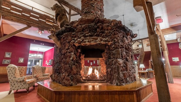 A stone fireplace with a fire, surrounded by wooden furniture and a red carpeted floor. - Heidelberg Inn (June Lake)