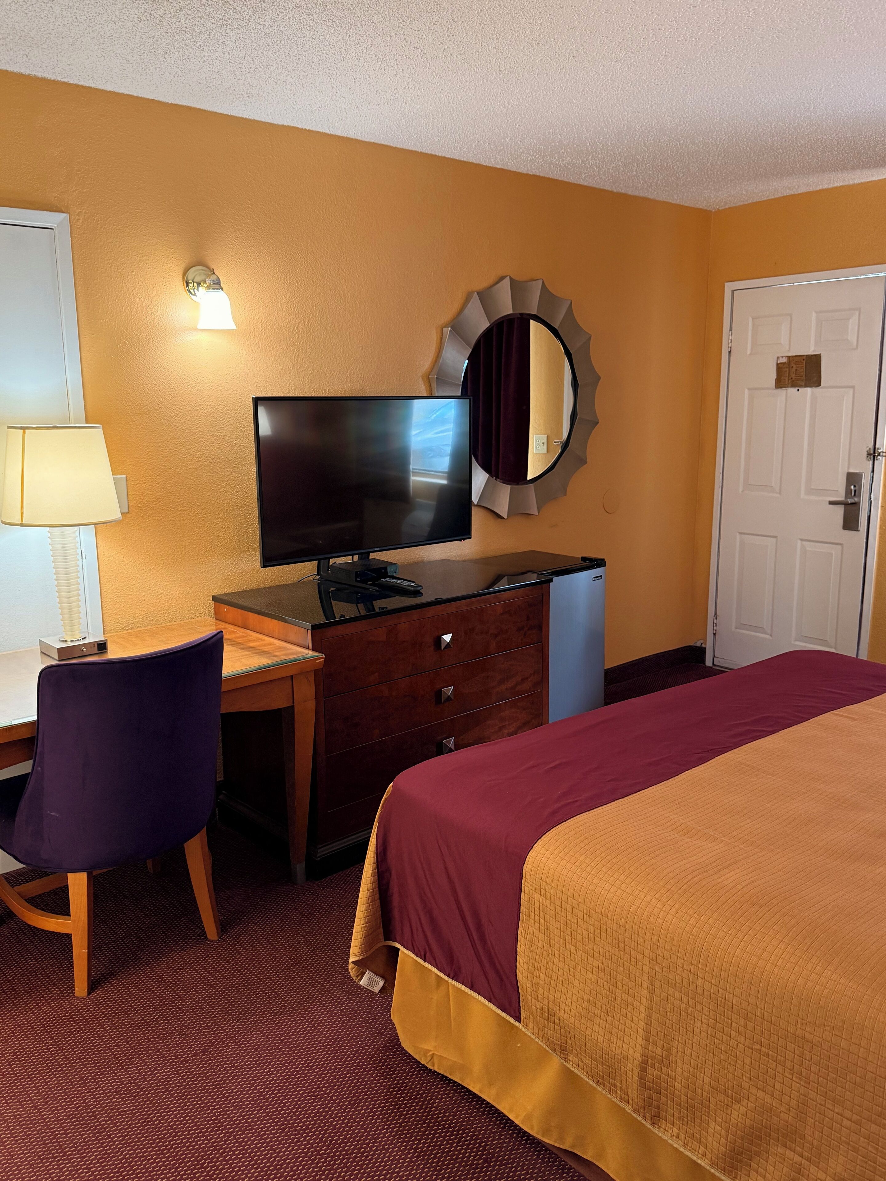 A hotel room with a bed, a desk with a chair, a television, a mirror, and a door.
