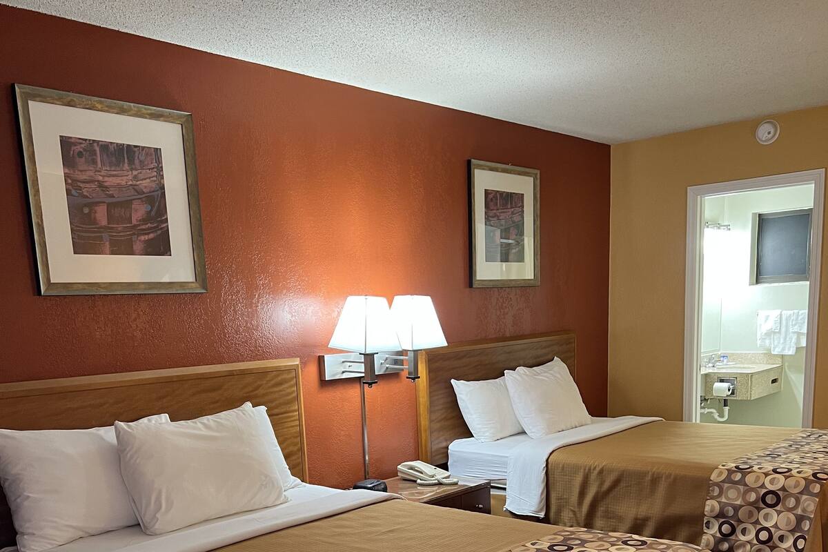 A hotel room with two beds, a nightstand, a lamp, and a bathroom visible through an open door.