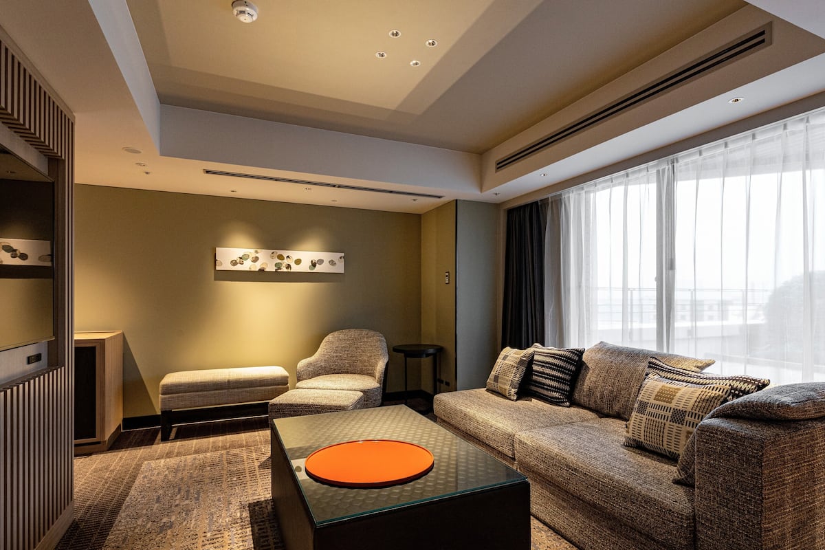 Une chambre d’hôtel moderne, dotée d’un canapé, d’un fauteuil et d’un pouf, d’une grande fenêtre avec des rideaux et d’une œuvre d’art accrochée au mur.