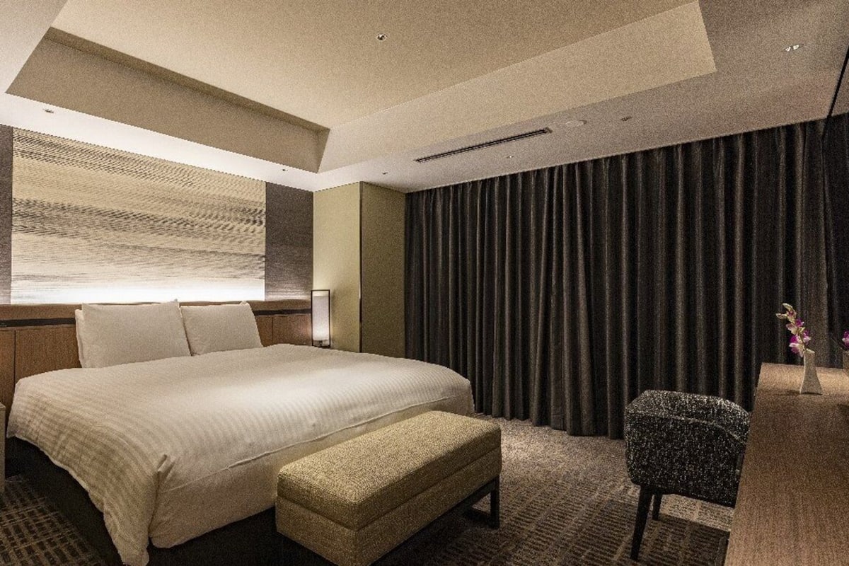 Une chambre d’hôtel moderne avec un grand lit, une banquette, une table de chevet et une lampe fixée au mur.