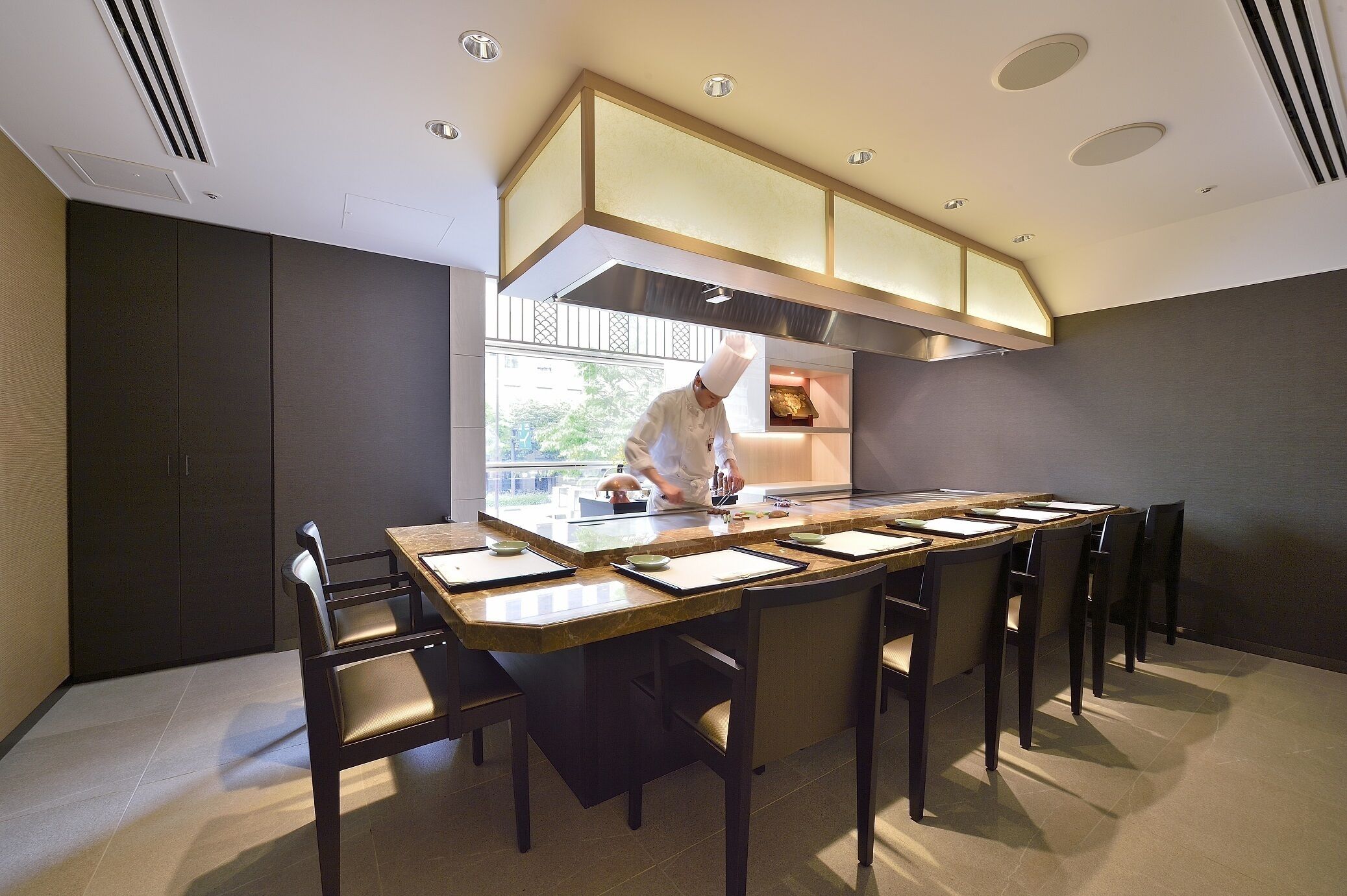 Una cocina moderna con un chef preparando comida, una larga mesa de comedor con sillas y un amplio ventanal que ofrece vistas al exterior.