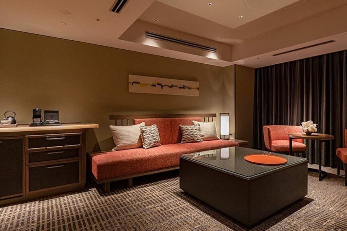 Une chambre d’hôtel moderne équipée d’un canapé, de deux fauteuils, d’une table basse, d’un bureau avec un ordinateur et d’une grande fenêtre avec des rideaux.