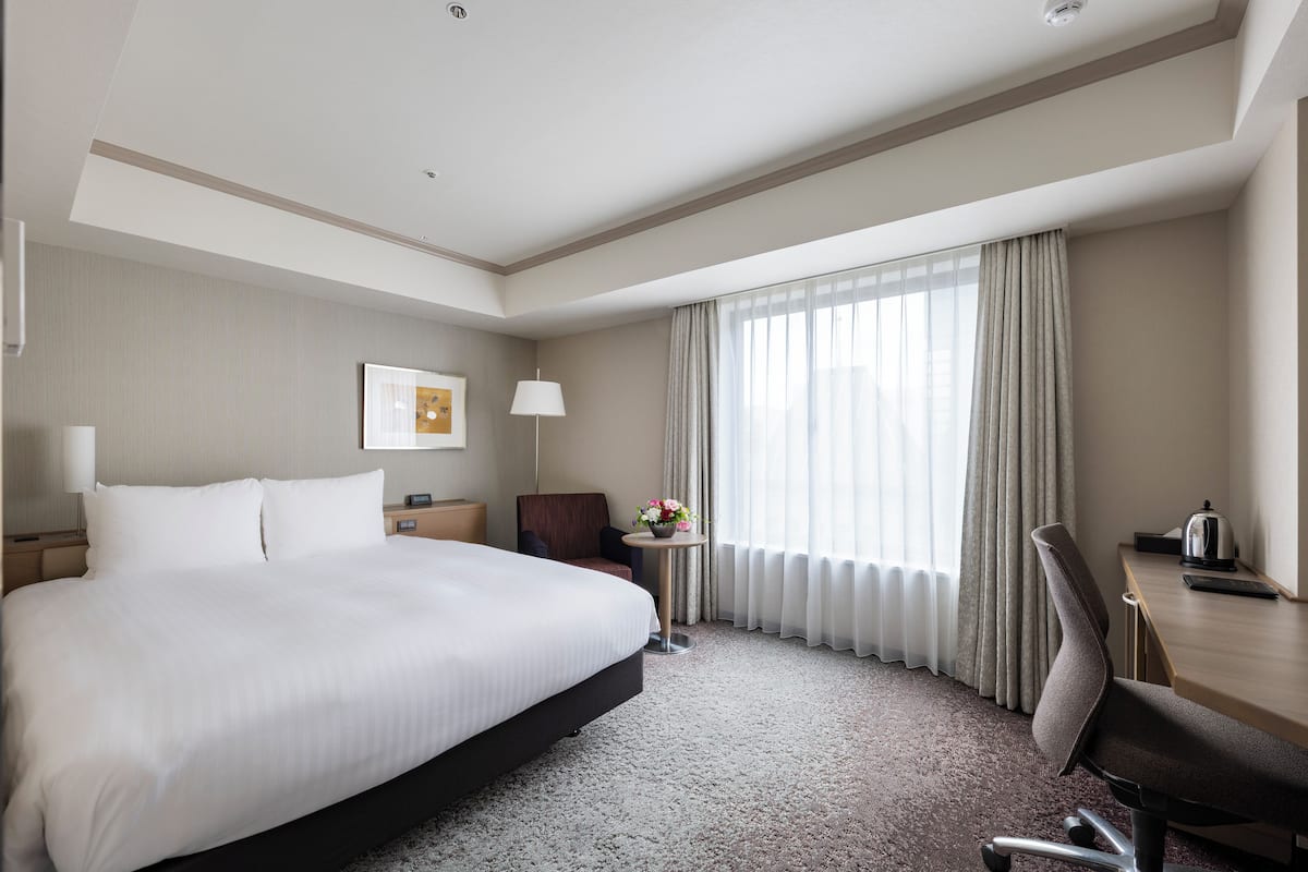 Une chambre d’hôtel avec un grand lit, un bureau, une chaise, une petite table avec une lampe et une fenêtre avec des rideaux.