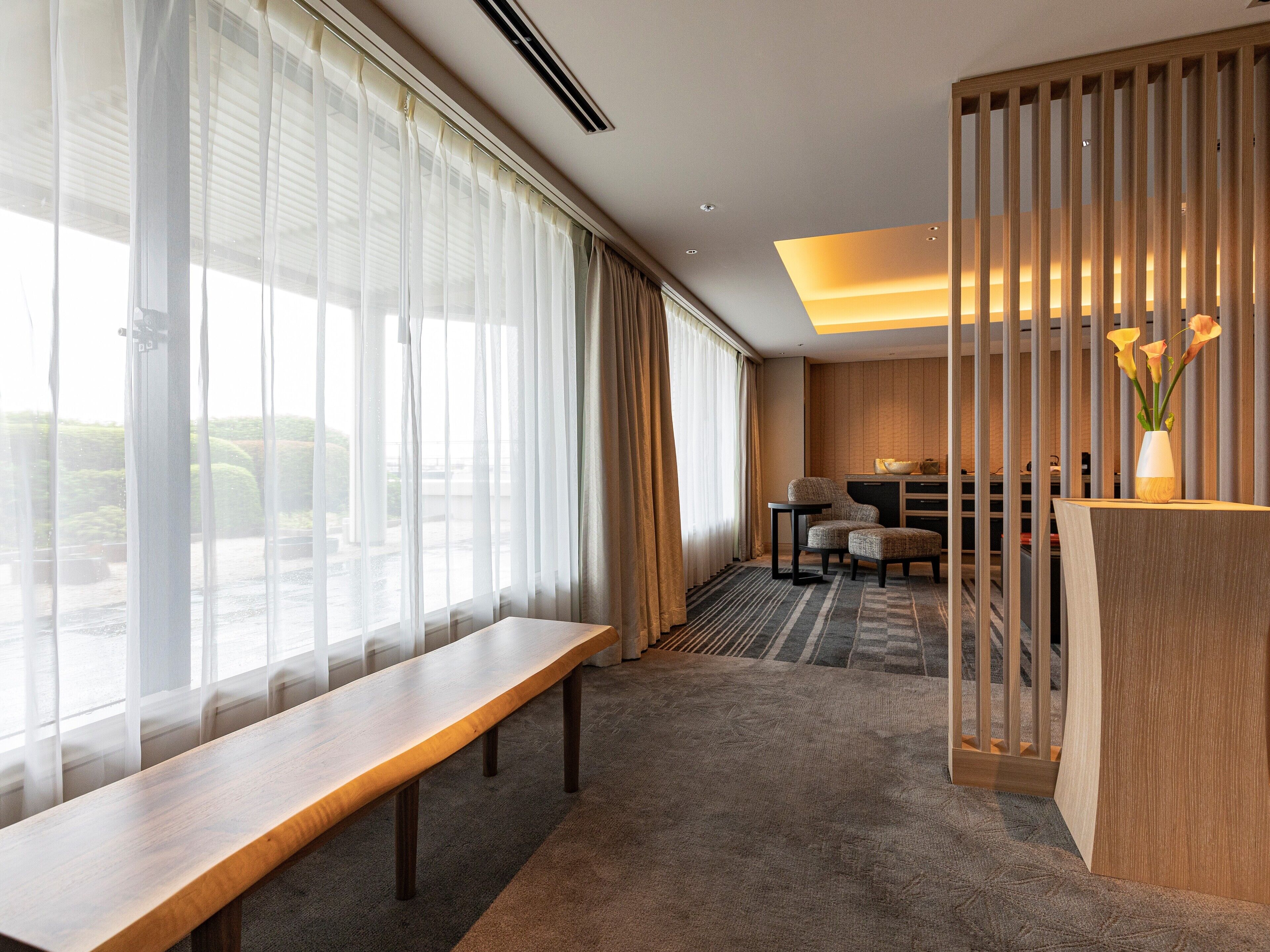 A hotel room with a large window, a wooden bench, a desk, and a partition with vertical slats.