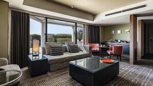 A modern hotel room with a sofa set, a glass coffee table, a small dining table, and a kitchen area.