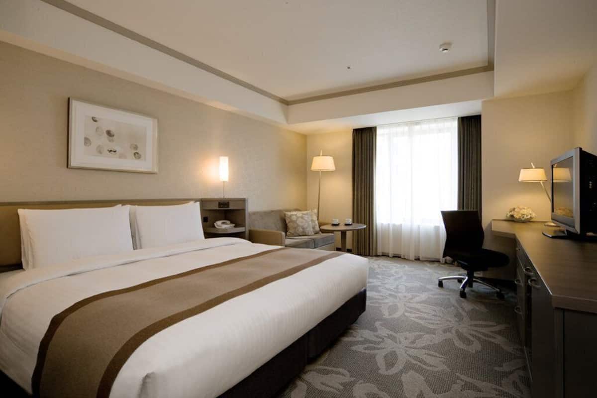 Une chambre d’hôtel avec un grand lit, un bureau avec un ordinateur, une chaise, une petite table et une fenêtre avec des rideaux.