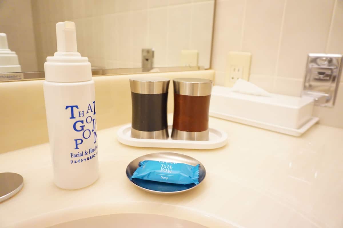Un lavabo de salle de bain avec un flacon de savon pour le visage et les mains, deux récipients métalliques et un porte-savon contenant un pain de savon.