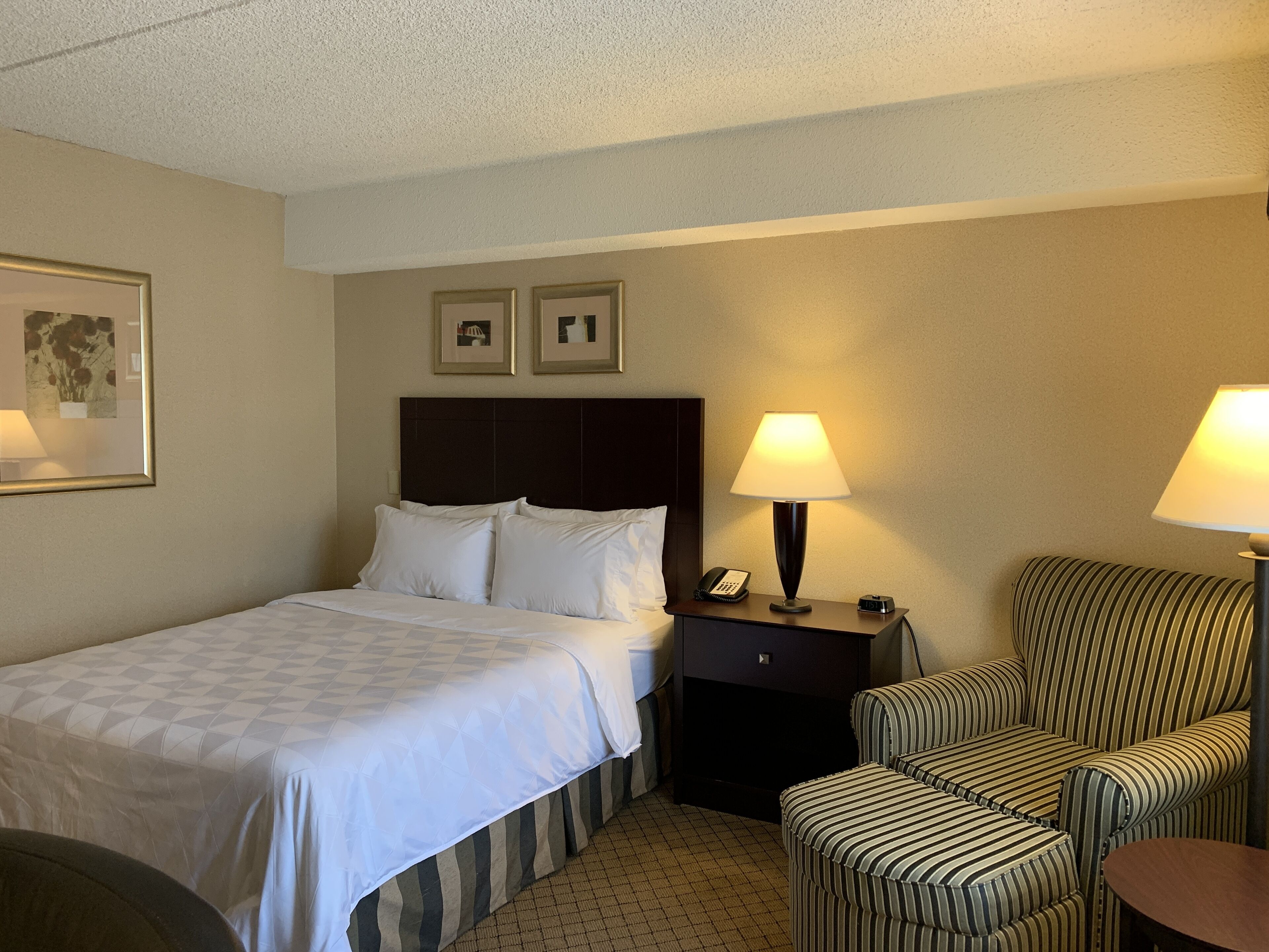 A hotel room with a bed, two bedside lamps, a nightstand, a striped armchair, and a framed picture on the wall.