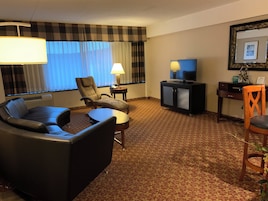 A hotel room with a sofa, armchair, a small table, a television, and a desk with a lamp.