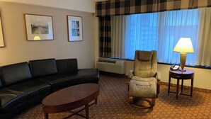 A hotel room with a black leather sofa, a wooden coffee table, a cushioned armchair, a standing lamp, and a window with curtains. - Holiday Inn Johnstown Downtown by IHG (Johnstown)