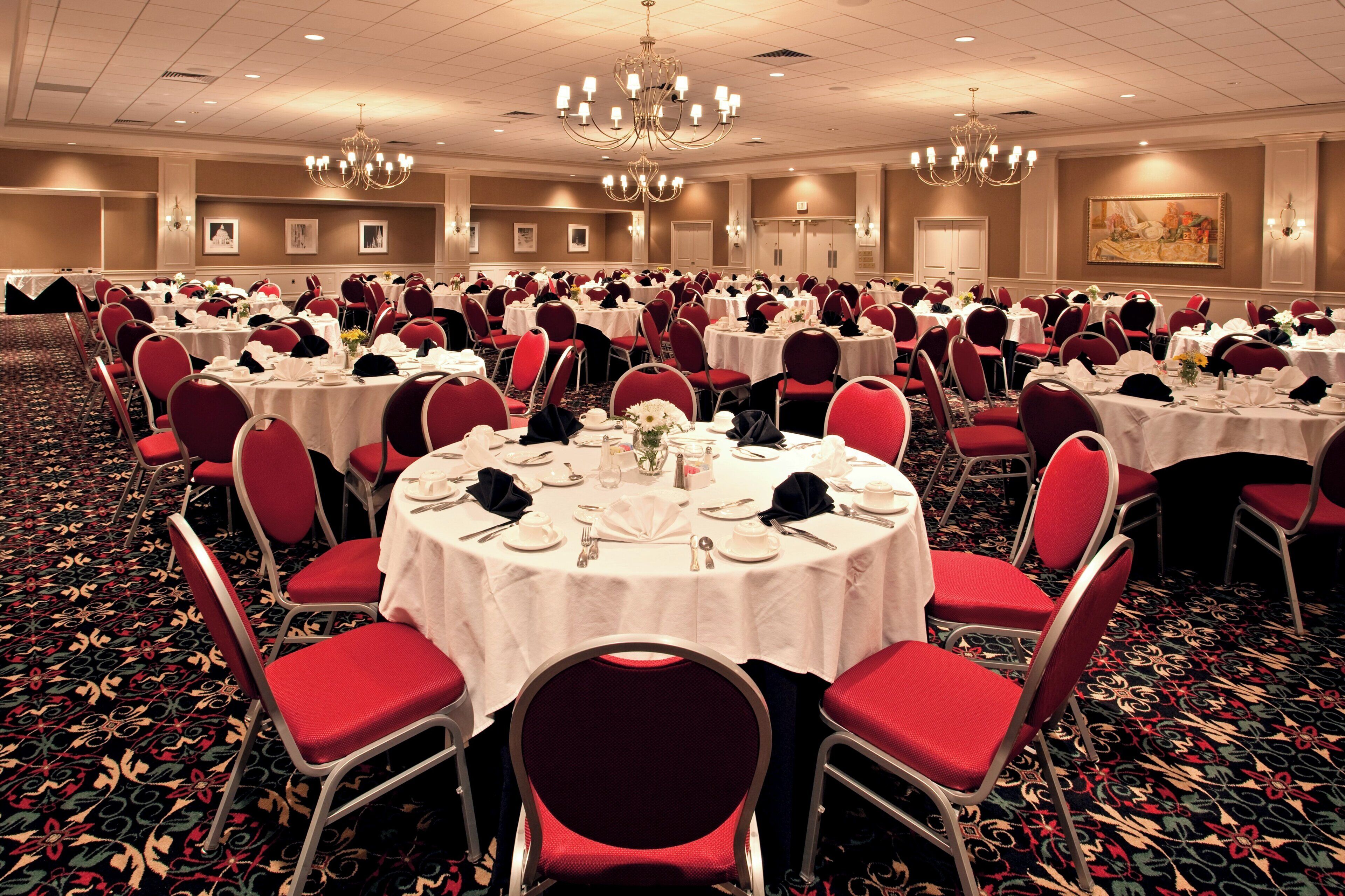 A banquet hall with round tables, red chairs, chandeliers, and a carpeted floor.