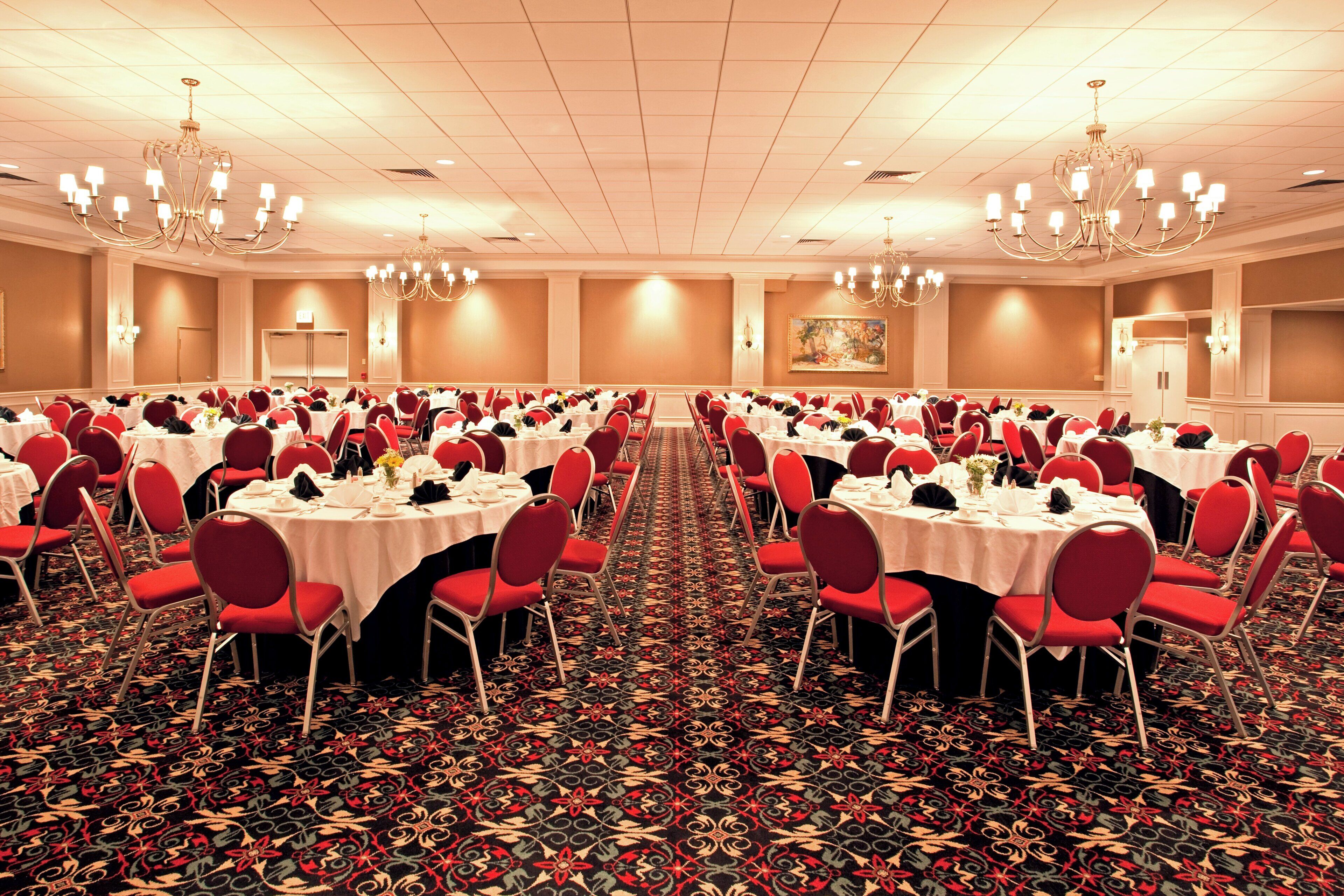 A banquet hall with round tables, red chairs, chandeliers, and a patterned carpet.