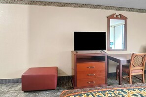 Habitación de hotel con cómoda de madera, televisión, espejo, reposapiés rojo y colcha estampada.
