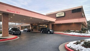 Un hotel con entrada accesible, un cartel que indica una altura libre de 3,8 metros, y un estacionamiento con varios automóviles.