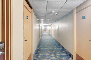 Pasillo de hotel con pisos alfombrados, puertas a ambos lados y un techo con cámara de seguridad.