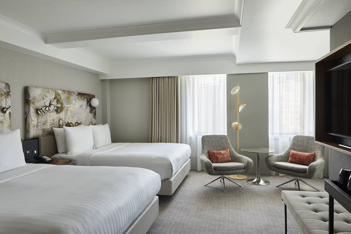 A hotel room with two beds, a TV, a desk, two armchairs, and a large window with curtains.