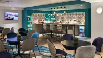 Un moderno hall de hotel con zona de bar, asientos y una pantalla de televisión que muestra un paisaje urbano.