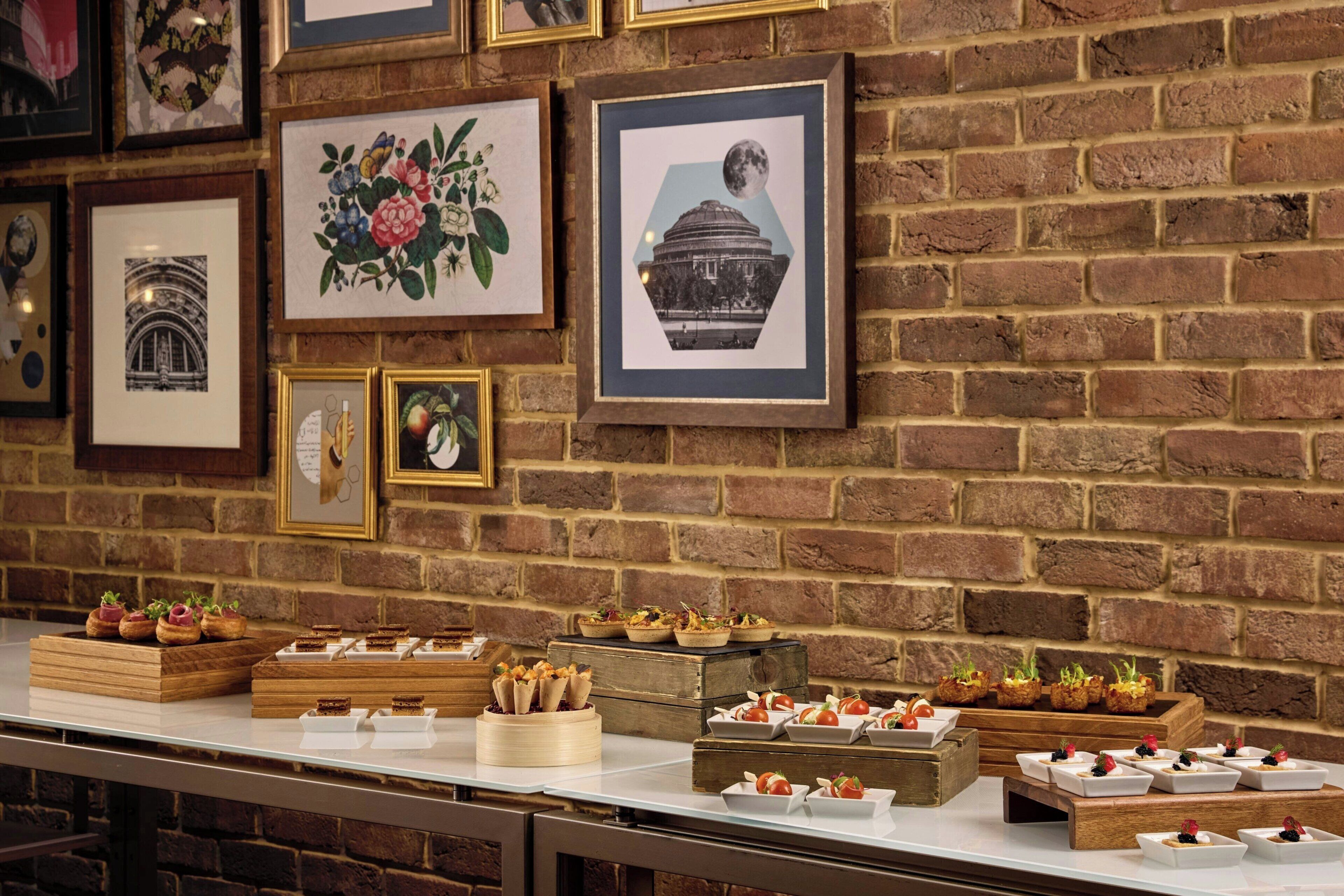 A buffet setup with various food items on trays against a brick wall with framed pictures.